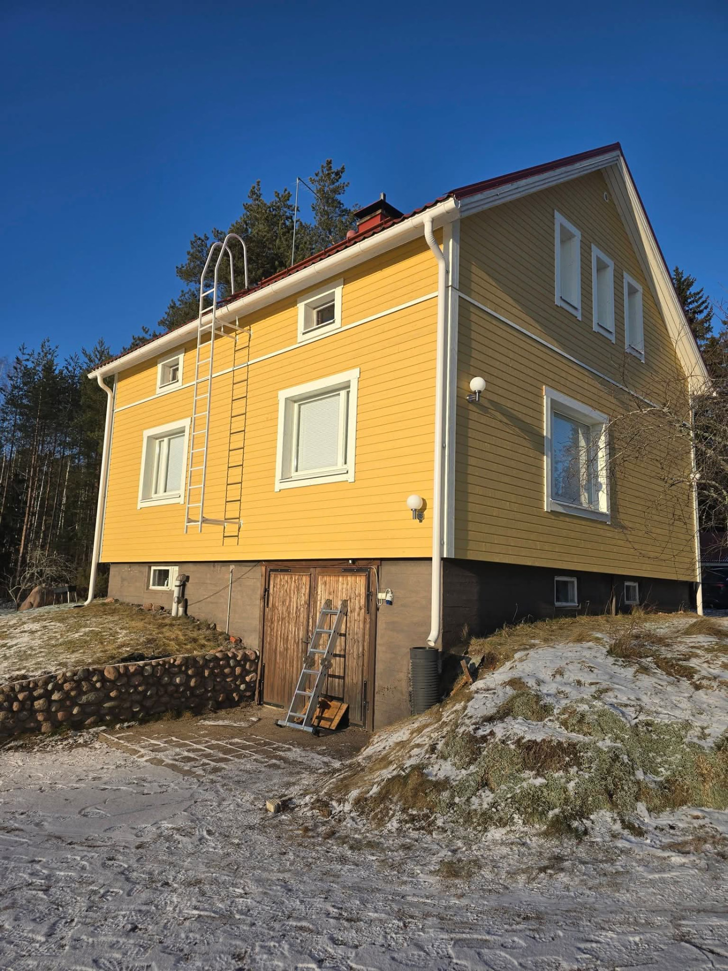 Keltainen puutalo harmaalla kivijalalla ja valkoisilla seinätikkailla