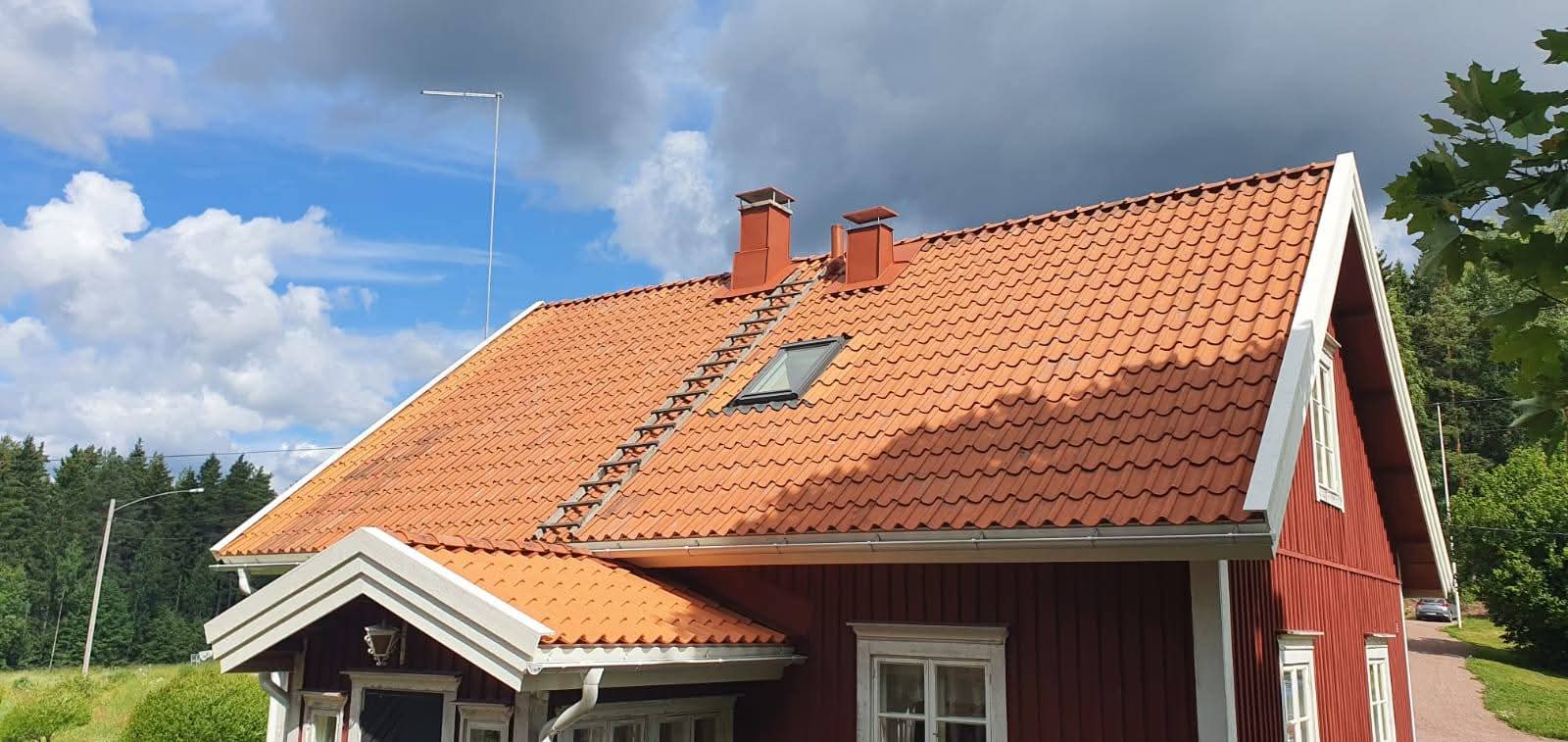 Punainen puutalo tiilikatolla ja valkoisilla listoituksilla, peltoja ja metsää taustalla